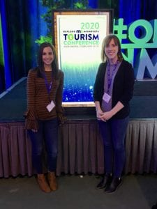 Two UMVRDC Staff posing in front of the podium that reads "2020 Explore Minnesota Tourism Conference."